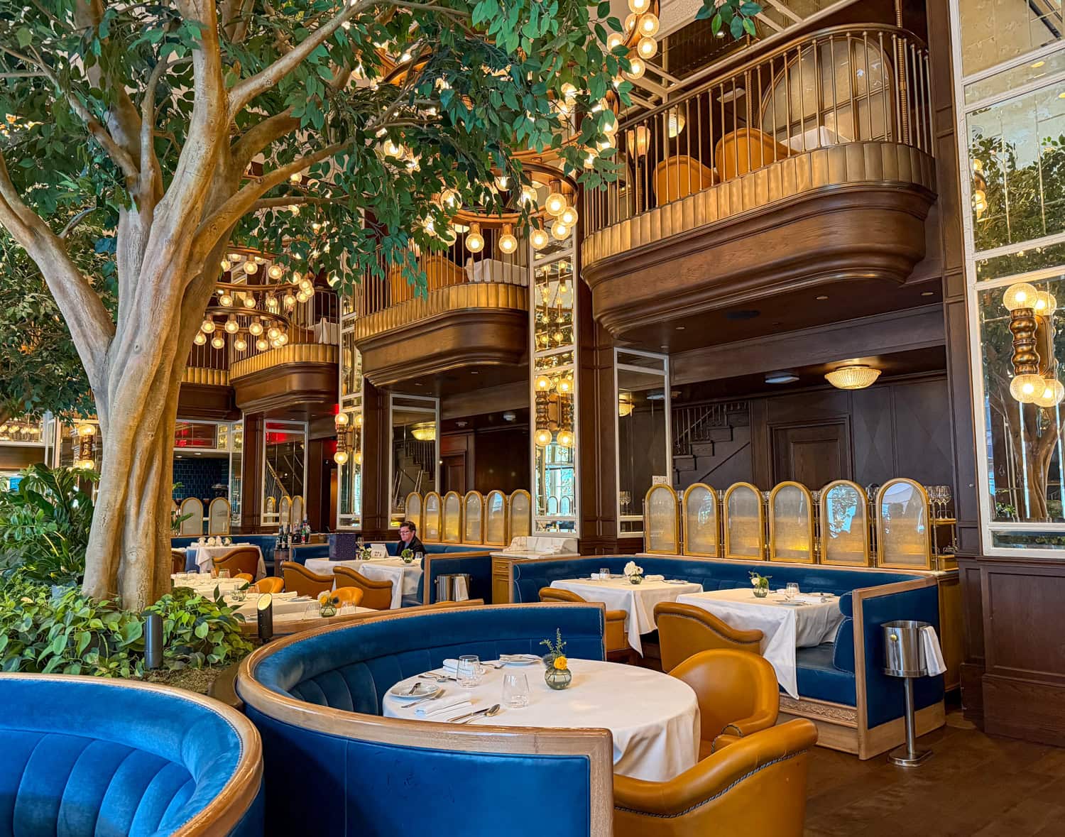 Elegant dining room at Café Carmellini in New York City with indoor trees, chandeliers, velvet booths, and warm lighting.