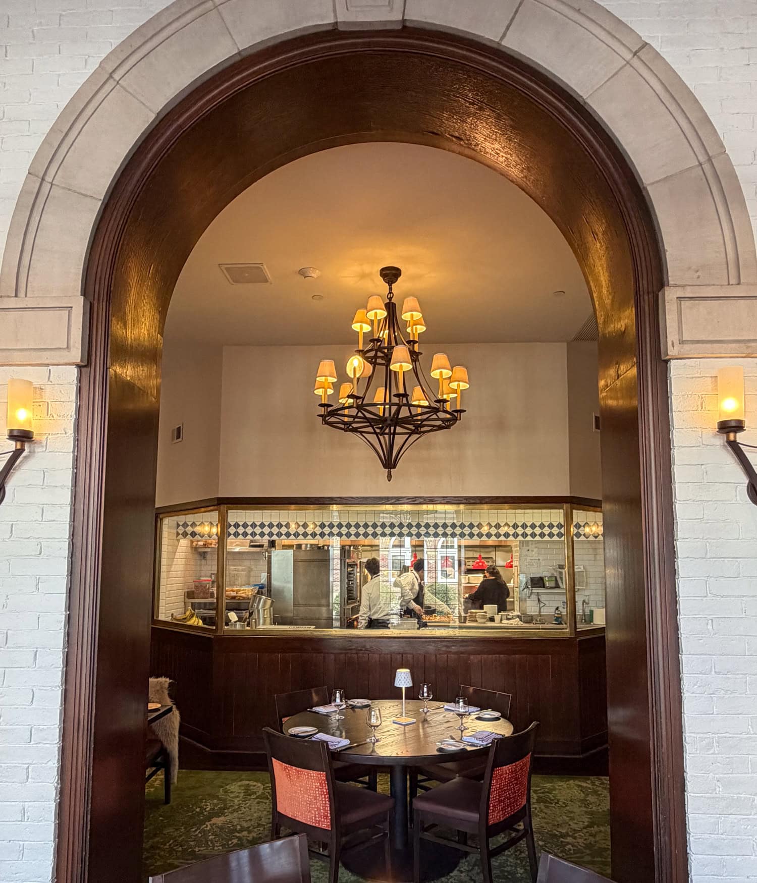 Dining table beneath an arch in American Bounty Restaurant, with CIA students working in the glass-enclosed pastry kitchen behind it.