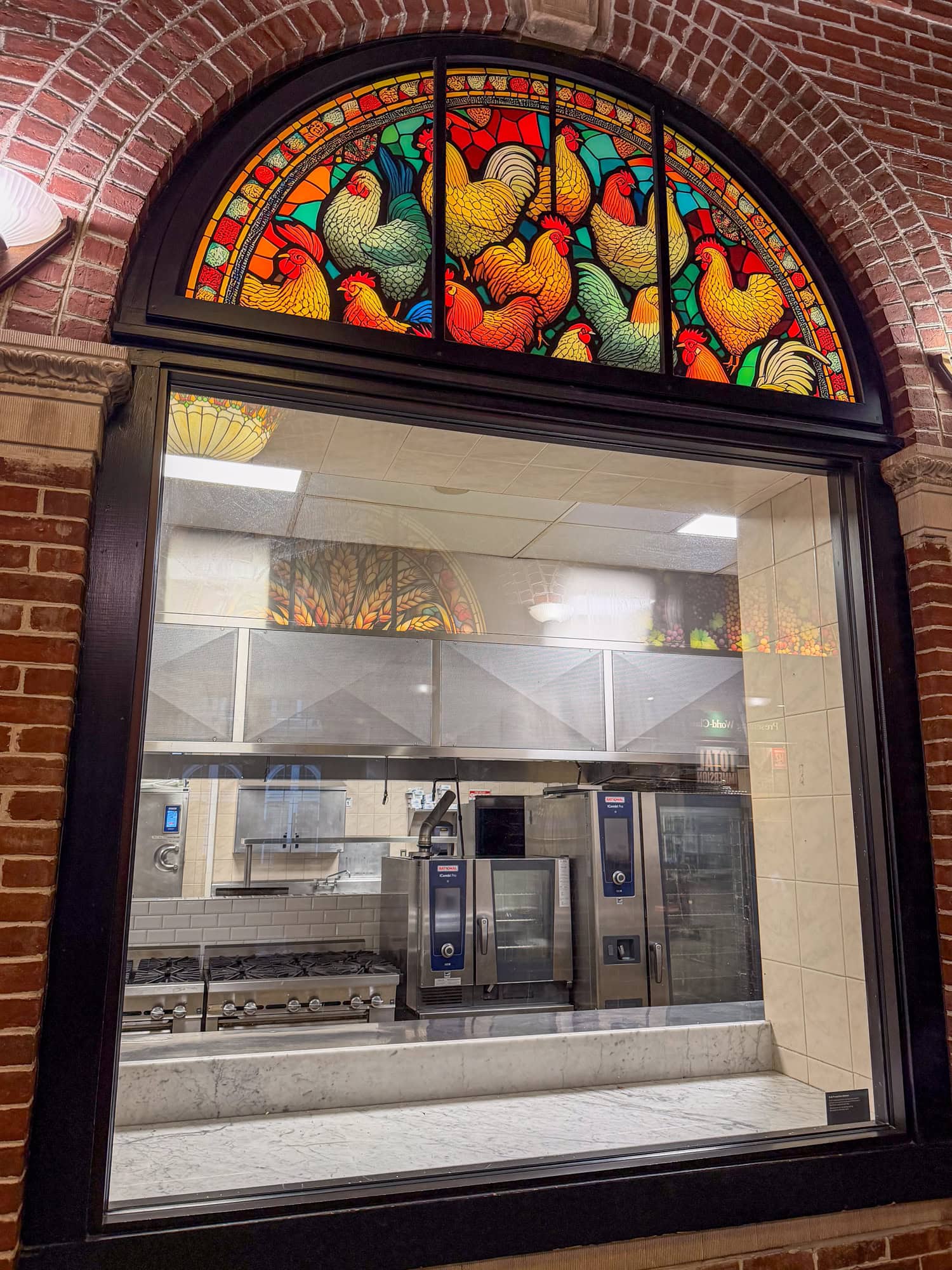 Empty CIA teaching kitchen visible through a window beneath stained glass depicting colorful chickens.
