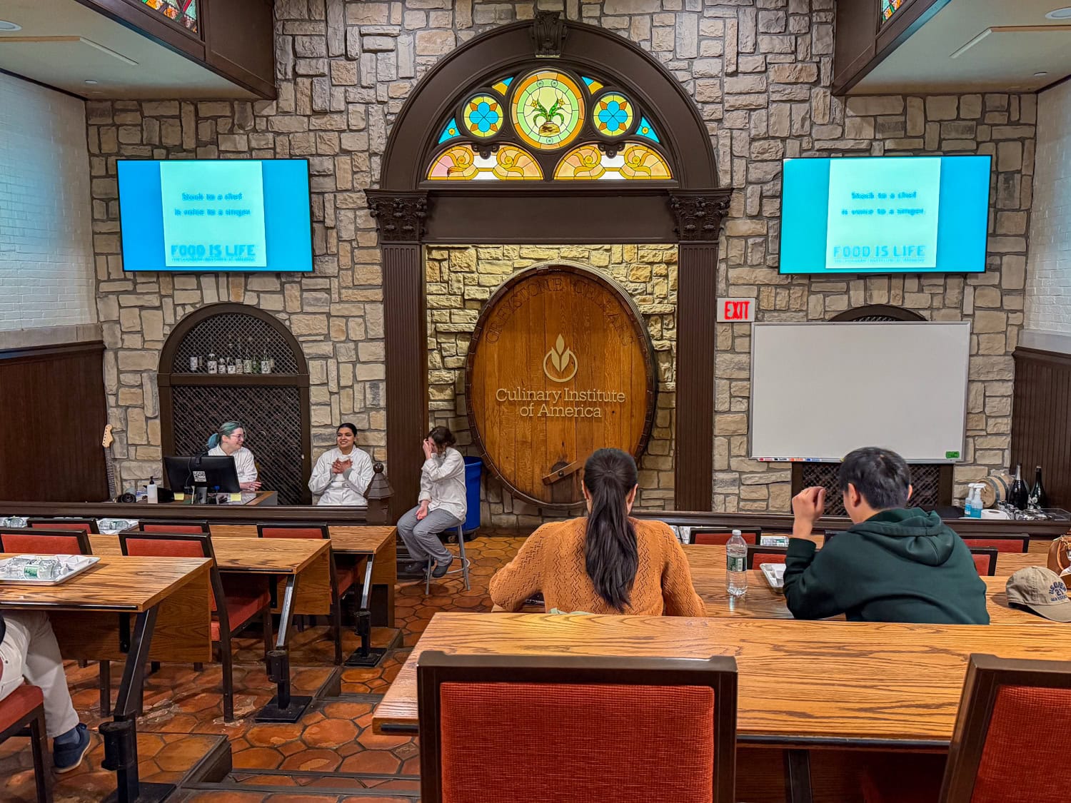 CIA tour participants seated in a stone-walled lecture hall with stained glass and a large wooden barrel displaying the school logo.