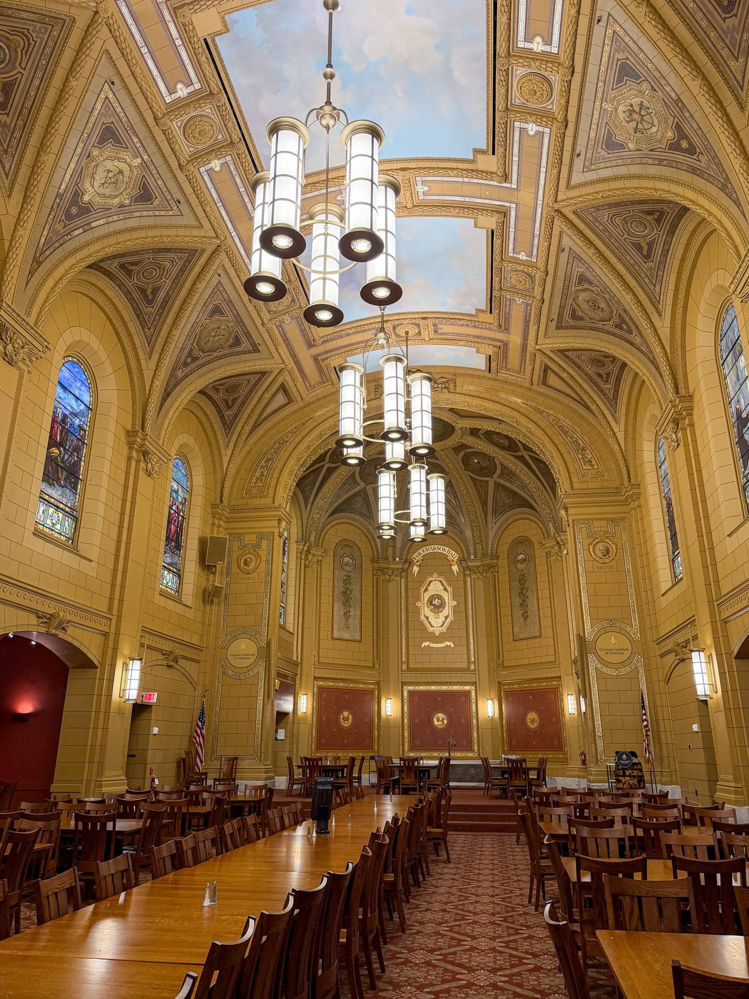 Farquharson Hall at the Culinary Institute of America, with a vaulted painted ceiling, chandeliers, stained-glass windows, and rows of wooden tables.