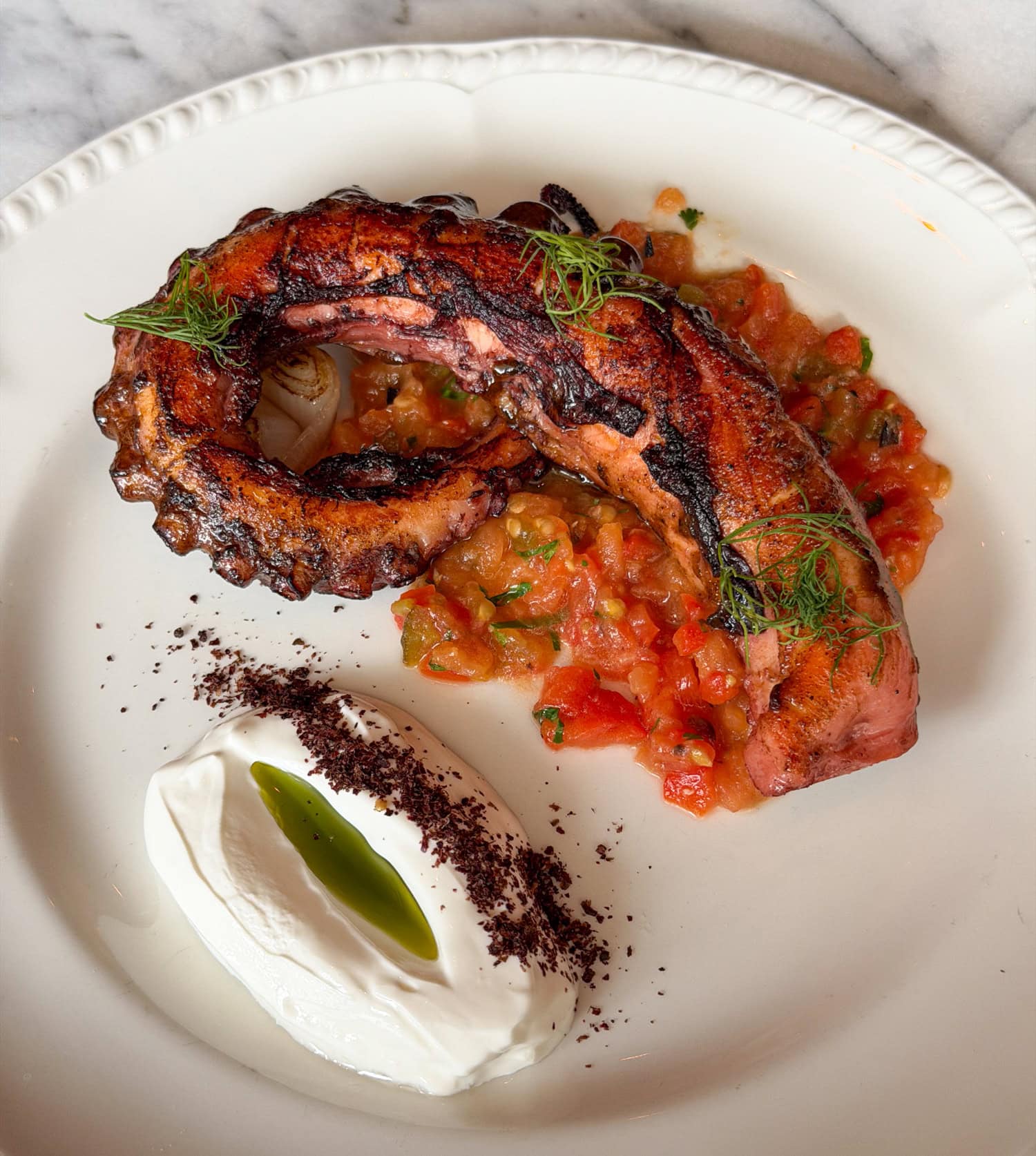 Charred octopus tentacle with herb garnishes, diced vegetables, and crème fraîche topped with green oil.