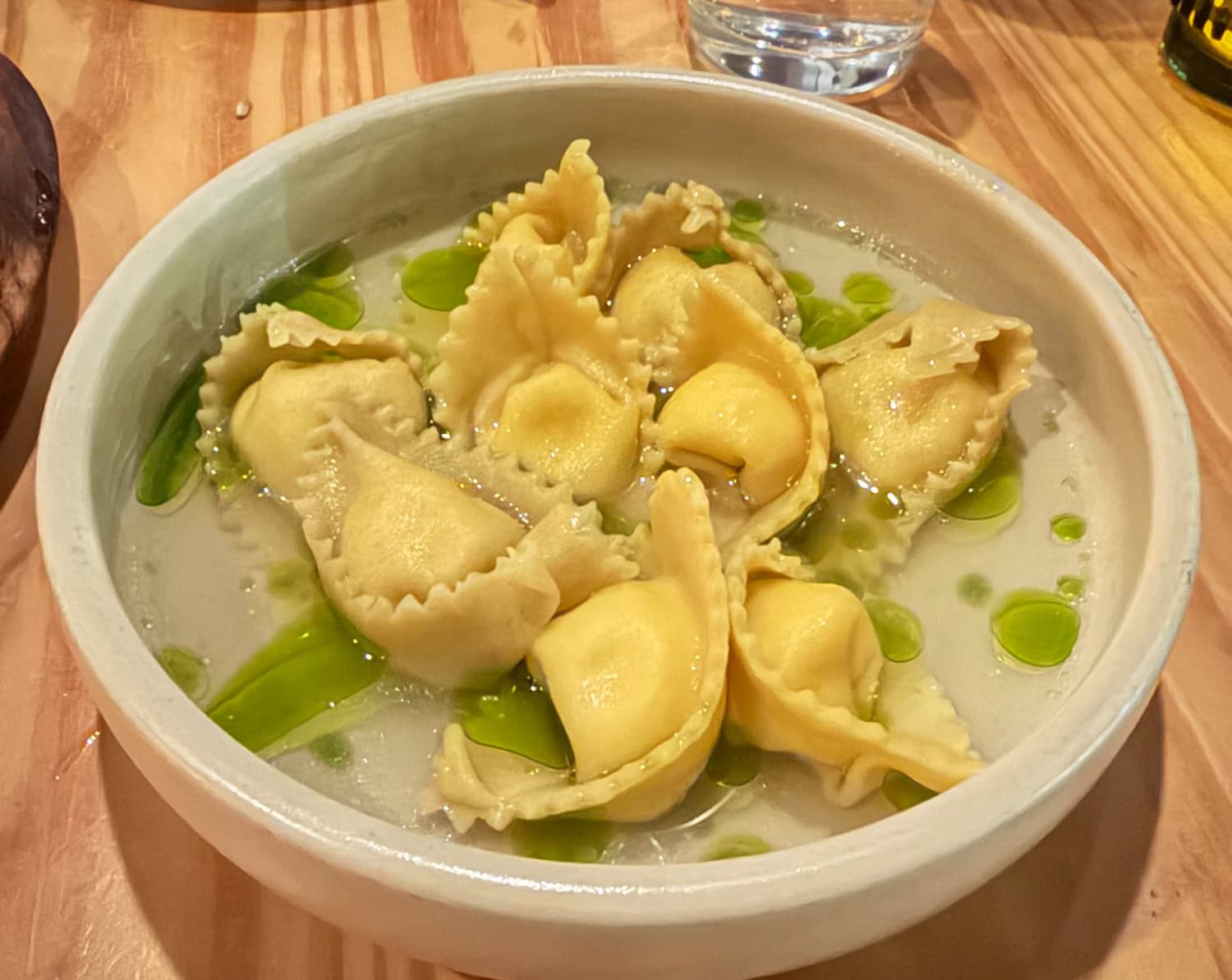 Folded gondolini pasta in a pale broth dotted with green chive oil, served in a ceramic bowl.
