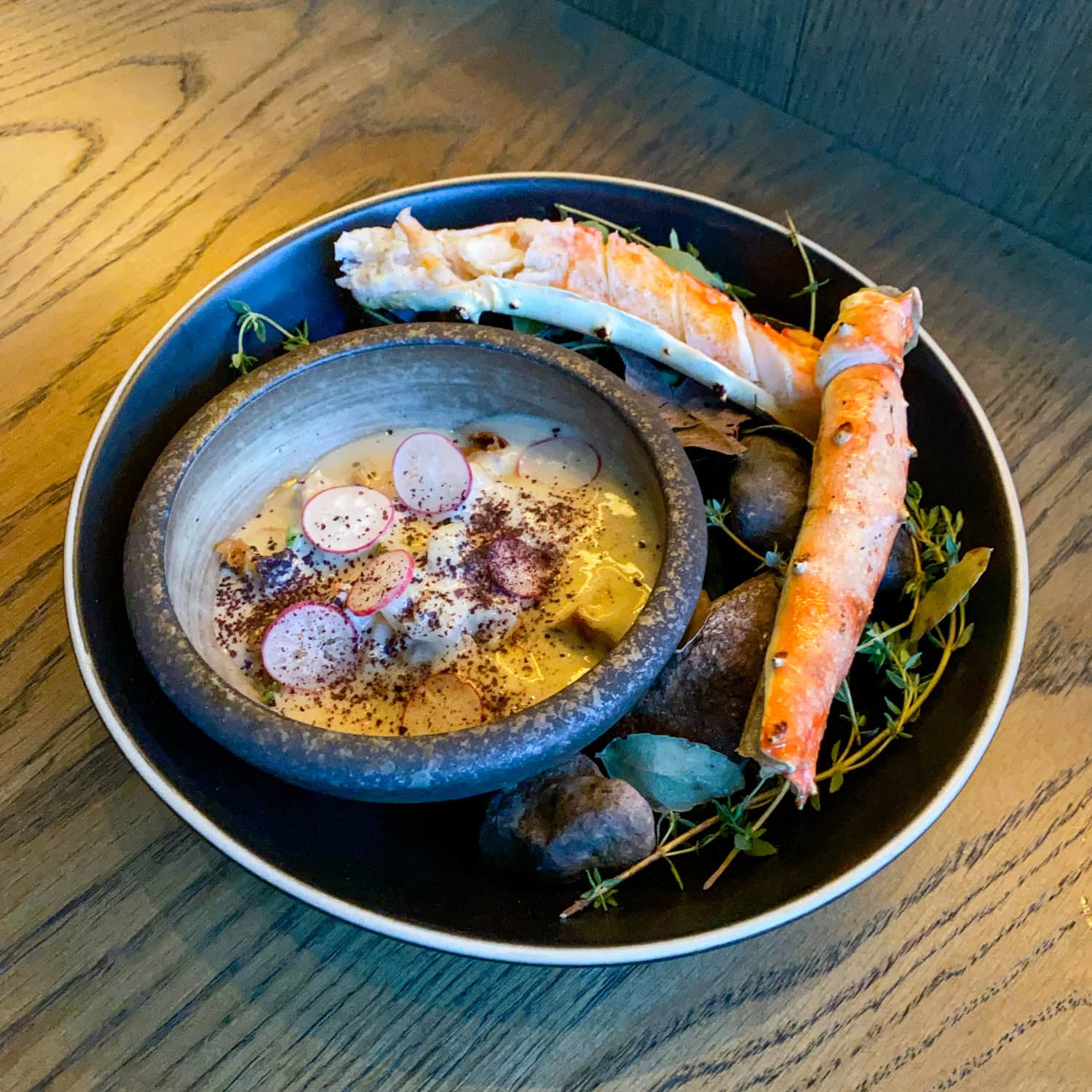 King crab presentation with orange shells, a ceramic bowl topped with sliced radishes, and herbs arranged on a dark platter.