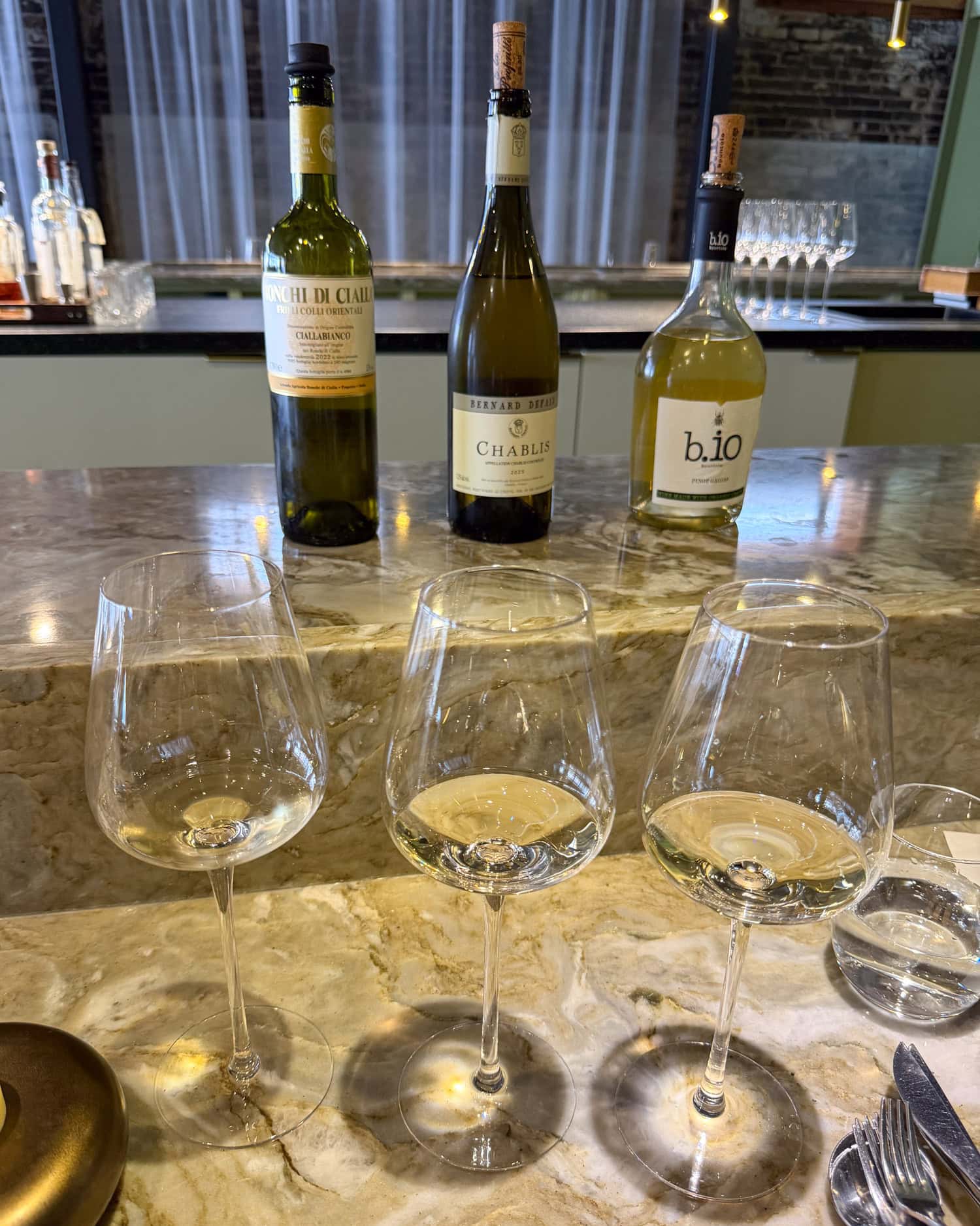 Three glasses of white wine samples lined up before their bottles on EBBE's marble counter.